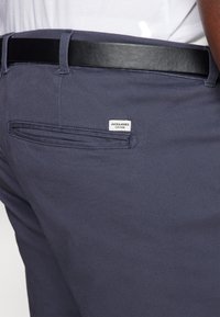 Dunkelblaue Hose mit glatter Stoffstruktur, ausgestattet mit einem schwarzen Gürtel und einer Gesäßtasche mit einem kleinen weißen Etikett, auf dem "JACK&JONES" steht.