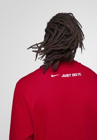 Červená mikina s texturovanou tkaninou a bílým logem "JUST DO IT" na zádech, s volným střihem a designem kulatého výstřihu.