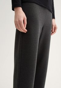 Divas rokas, kas atpūšas blakus cilvēka vēderam, kurš ģērbies tumši pelēkos adījumos un melnā garā piedurknē, pret vienkāršu fonu.