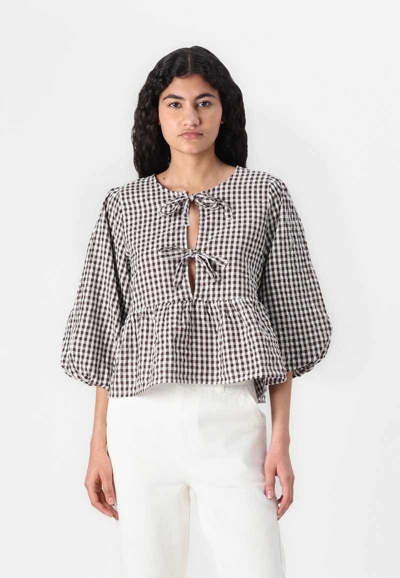 Blusa a quadri marrone e bianca con maniche a palloncino, scollatura a chiave con chiusura a lacci e dettaglio peplum, abbinata a pantaloni bianchi.