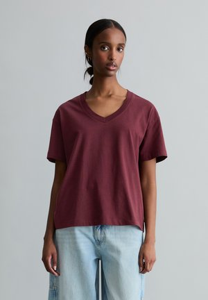 Junge Frau mit geflochtenen Haaren trägt ein locker sitzendes, bordeauxrotes T-Shirt mit V-Ausschnitt und hellblaue, weit geschnittene Jeans vor einem schlichten grauen Hintergrund.