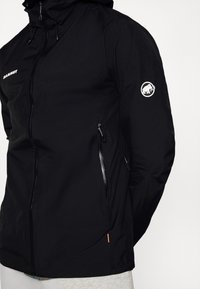 Veste noire à capuche avec fermeture éclair frontale, poches latérales zippées et logo mammouth blanc sur la partie supérieure de la manche.