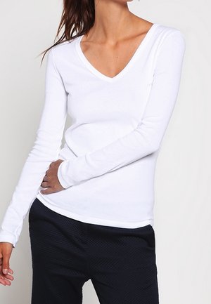 Vrouw draagt een wit, nauwsluitend, V-hals shirt met lange mouwen en textuurrijke zwarte broek, poseert met één hand op de heup tegen een neutrale achtergrond.