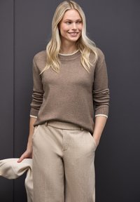 Brązowy sweter wełniany z prążkowanymi mankietami i dekoltem, w połączeniu z jasnobeżowymi, teksturowanymi spodniami. Model trzyma jasnobeżową kurtkę.