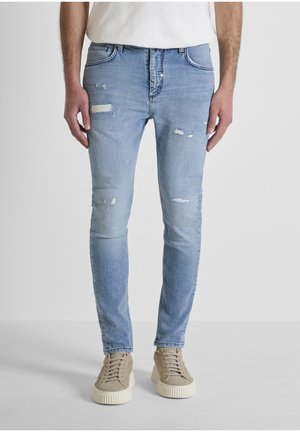 Persona vistiendo jeans ajustados desgastados de color azul claro con pequeños desgarros, camiseta blanca y zapatillas beige, de pie sobre un suelo gris.