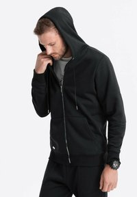 Sort lynlås hoodie med et struktureret stof, stor hætte og snoretræk. Har frontlommer og en lille logo detalje.