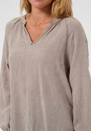 Femme portant un chemisier beige clair texturé avec des motifs floraux brodés et un collier en or avec deux petites bagues entrelacées.