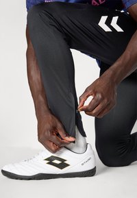 Pantalons de sport noirs avec une cheville zippée, assortis à des chaussures de sport blanches et noires, arborant un logo et un design texturé.