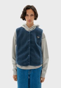 Niebieska kamizelka polarowa o miękkiej fakturze, z zatrzaskami i naszywką z logo, noszona na szarym hoodie i niebieskich dżinsach. Prosty, casualowy design.