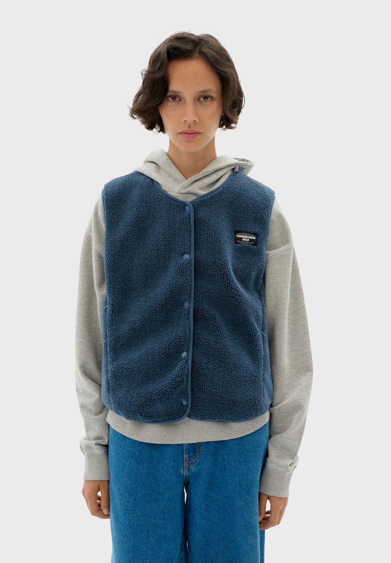 Niebieska kamizelka polarowa o miękkiej fakturze, z zatrzaskami i naszywką z logo, noszona na szarym hoodie i niebieskich dżinsach. Prosty, casualowy design.