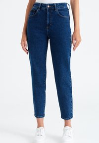 Džínové kalhoty s vysokým pasem v modré barvě, rovný střih nohavic, pět kapes a jemné vyblednutí. Doplněno bílými teniskami na šněrování.