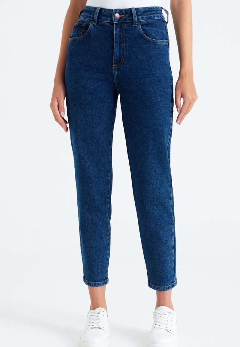 Džínové kalhoty s vysokým pasem v modré barvě, rovný střih nohavic, pět kapes a jemné vyblednutí. Doplněno bílými teniskami na šněrování.