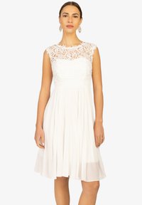 Robe blanche avec un corsage en dentelle et une jupe plissée. Modèle sans manches avec un col haut et des détails en dentelle florale complexe en haut.