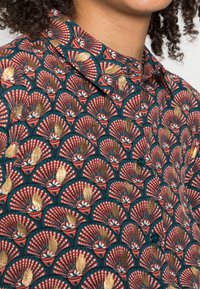 Chemise boutonnée avec un fond turquoise et un motif de ventilateur en rouge, blanc et or. Tissu léger avec un col et des boutons.