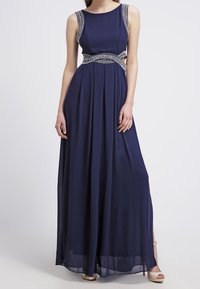 Navy-blaues ärmelloses Abendkleid mit silbernen Perlendetails an den Trägern und der Taille, getragen mit beigen High Heels.