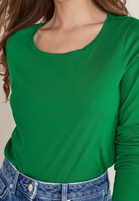 Green long-sleeve top with a scoop neckline, made of soft fabric. Paired with high-waisted blue denim jeans.