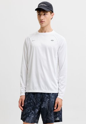 Jeune homme portant une chemise blanche à manches longues, un short noir à motifs et une casquette bleu marine sur un fond clair uni.