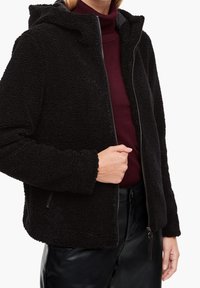 Giacca in pile nera con zip frontale, cappuccio e tessuto testurizzato. Presenta polsini elasticizzati e un orlo aderente. Indossata su un dolcevita bordeaux.