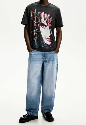 Osoba ubrana w czarną koszulkę z grafiką twarzy i tekstem, luźne jasnoniebieskie dżinsy i czarne buty, stojąca z rękami w kieszeniach.