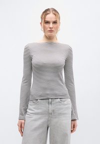 Ριγέ μπλούζα με μακριά μανίκια σε γκρι και λευκό βαμβάκι, με λαιμόκοψη καραβόπανο και στενή γραμμή. Φορεμένη με τζιν σε ανοιχτό γκρι.