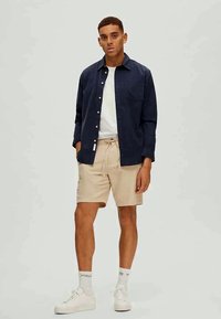 Tengeri gombos ing egy fehér póló felett, bézs rövidnadrággal és fehér sportcipővel párosítva. Simára kidolgozott anyag, laza fazon és minimális kiegészítők.