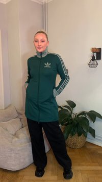 Casaco verde com fecho de correr, mangas compridas e logótipo adidas, combinado com calças pretas. Apresenta detalhes cinzentos contrastantes e dois bolsos laterais.