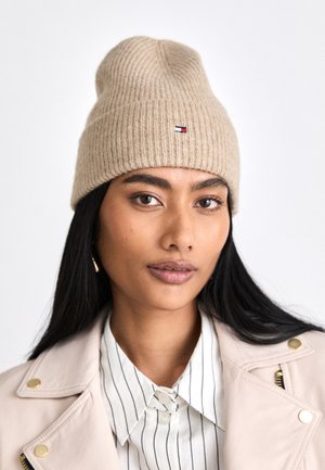Femme aux longs cheveux noirs portant un bonnet en tricot beige, une chemise rayée à col et une veste claire avec des boutons dorés.