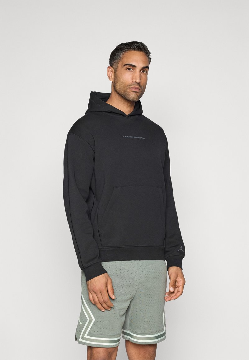 Μαύρη μπλούζα hoodie με τσέπη καγκουρό και διακριτό λογότυπο στο στήθος; ο χρήστης έχει συνδυάσει με ανοιχτό πράσινες διάτρητες βερμούδες με ρίγες.