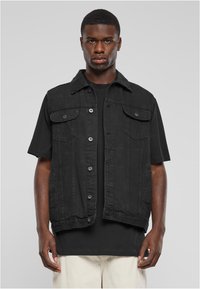 Joven vistiendo un chaleco de mezclilla sin mangas negro abotonado sobre una camiseta negra y pantalones de color claro, frente a un fondo pálido.