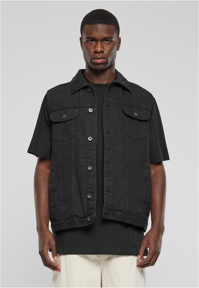 Joven vistiendo un chaleco de mezclilla sin mangas negro abotonado sobre una camiseta negra y pantalones de color claro, frente a un fondo pálido.