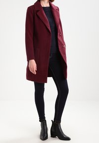 Manteau en mélange de laine bordeaux avec revers crantés, boutons croisés et poches latérales. Porté avec un jean sombre ajusté et des bottines noires.