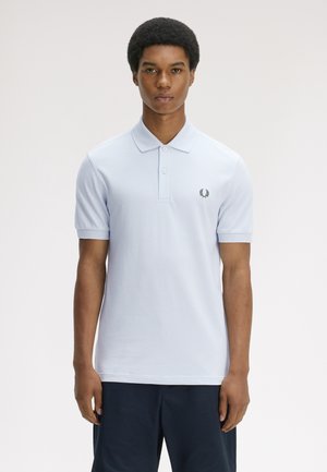Ung mand med kort krøllet hår iført lyseblå polo-shirt og mørke shorts, stående mod en ensfarvet lys baggrund.