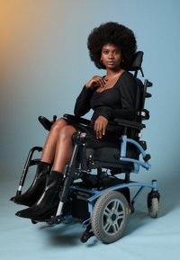 Νεαρή γυναίκα με αφρικάνικο μαλλί, ντυμένη με μαύρο φόρεμα και μπότες, κάθεται με αυτοπεποίθηση σε ένα μπλε ηλεκτρικό αναπηρικό καροτσάκι με φόντο απλή επιφάνεια.