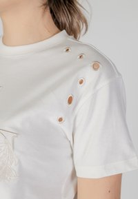 White cotton t-shirt with round cut-outs on one shoulder, featuring beige embroidery on the front and a soft, smooth texture.