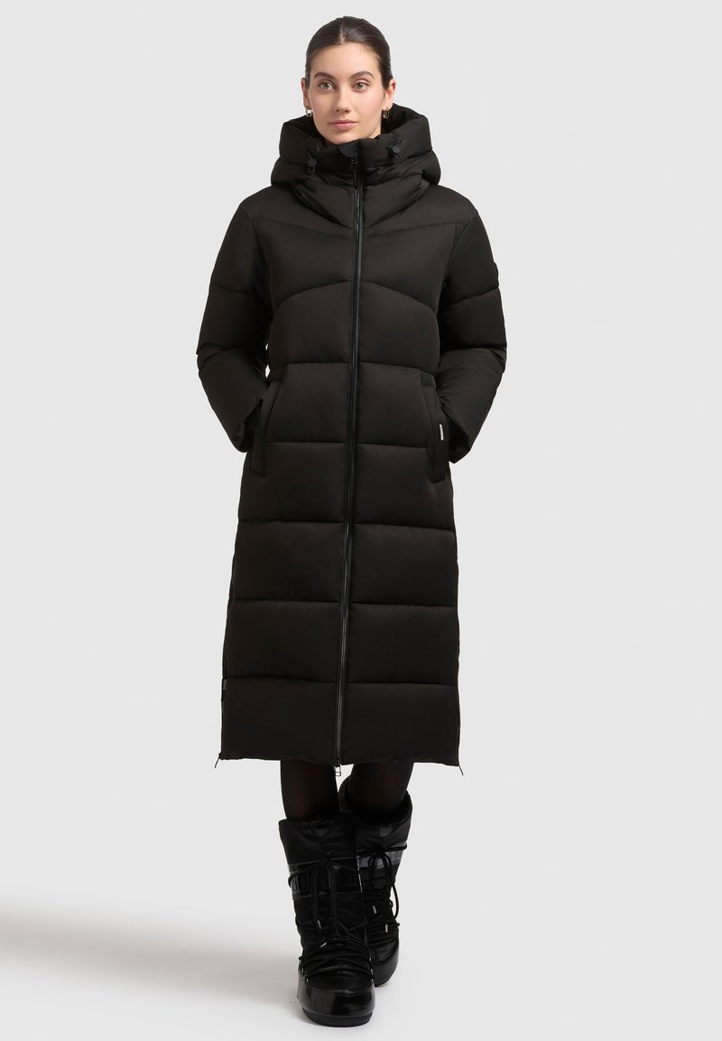 Musta pitkä toppatakki hupulla, jossa on edessä vetoketju, sivutaskut ja tikattu kuosi eristyksen ja lämmön takaamiseksi.