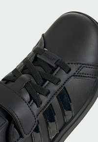 Black athletic shoe with a textured leather upper, glossy black accents, laces, and a strap. Features three black-striped branding on the side.
