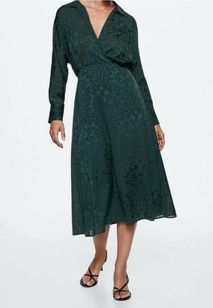 Mujer con un vestido midi verde oscuro de manga larga con sutiles estampados florales y tacones negros de tiras y punta abierta.