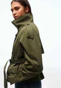 Giacca verde oliva con colletto alto, design corto e vita con cintura. Presenta dettagli con bottoni e spalline. Tessuto dalla texture liscia.