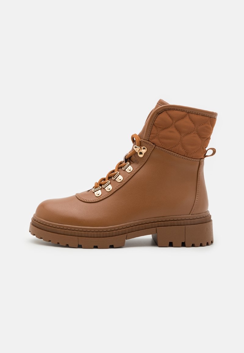 Ruskea nilkkaboots, joka on valmistettu synteettisestä materiaalista. Bootseissa on tikattu päällinen, pyöreä kärki, kultasävytetyt rei'itykset ja teksturoitu pohja.