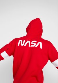 Červená mikina s kapucí s velkým bílým logem „NASA“ na zádech, s bílými detaily na rukávech a stahovací kapucí. Měkká texturovaná látka.