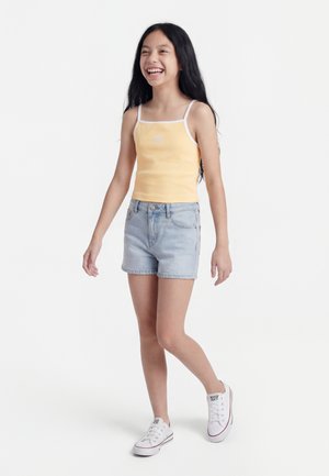 Dievča s dlhými tmavými vlasmi, usmievajúce sa, oblečené v žltom tielku bez rukávov, svetlom rifľovom šortkách a bielych teniskách, kráčajúce na bielom pozadí.