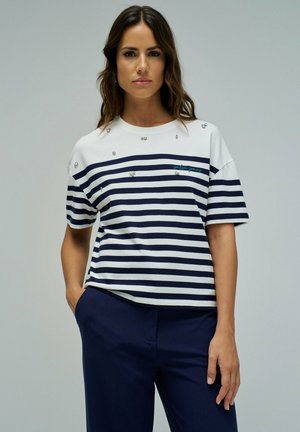 Femme portant une chemise blanche à rayures horizontales bleu marine avec de petites décoration, associée à un pantalon bleu foncé, debout avec une main dans la poche.