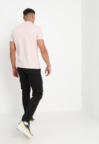 Jasnoróżowa koszulka polo, czarne spodnie cargo i białe sneakersy z żółtymi akcentami. Ujęcie z tyłu, pokazujące szczegóły swobodnego stroju.