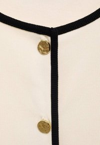 Vêtement crème à boutons avec un col et des bordures noir côtelé, présentant un décolleté rond et un bouton doré brillant à l'avant.