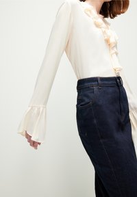 Frau in einer cremefarbenen Bluse mit Rüschen und weit ausgestellten Ärmeln, kombiniert mit einem hoch sitzenden, dunklen Jeansrock vor einem schlichten Hintergrund.