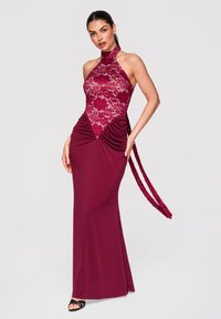 Femme portant une robe bordeaux sans manches avec un corsage en dentelle, un col halter, une taille froncée et de longues bretelles nouées, posant devant un fond uni.