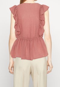 Roséblus med volanger och en strukturerad, randig design, med en peplumslutning och genomskinligt tyg. Bakifrån fokuserar bilden på volangerna.