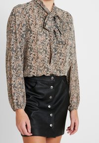 Blusa a maniche lunghe in tessuto fantasia marrone, beige e nero con un fiocco al collo, abbinata a una mini gonna nera in pelle con bottoni frontali.