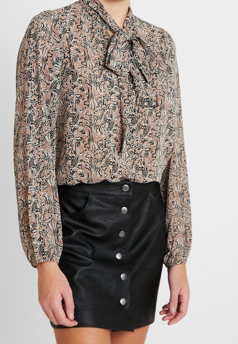 Blusa a maniche lunghe in tessuto fantasia marrone, beige e nero con un fiocco al collo, abbinata a una mini gonna nera in pelle con bottoni frontali.