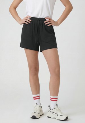 Persona in piedi con le mani sui fianchi che indossa pantaloncini neri con coulisse, scarpe da ginnastica bianche, calzini bianchi con strisce rosse e una maglietta bianca.